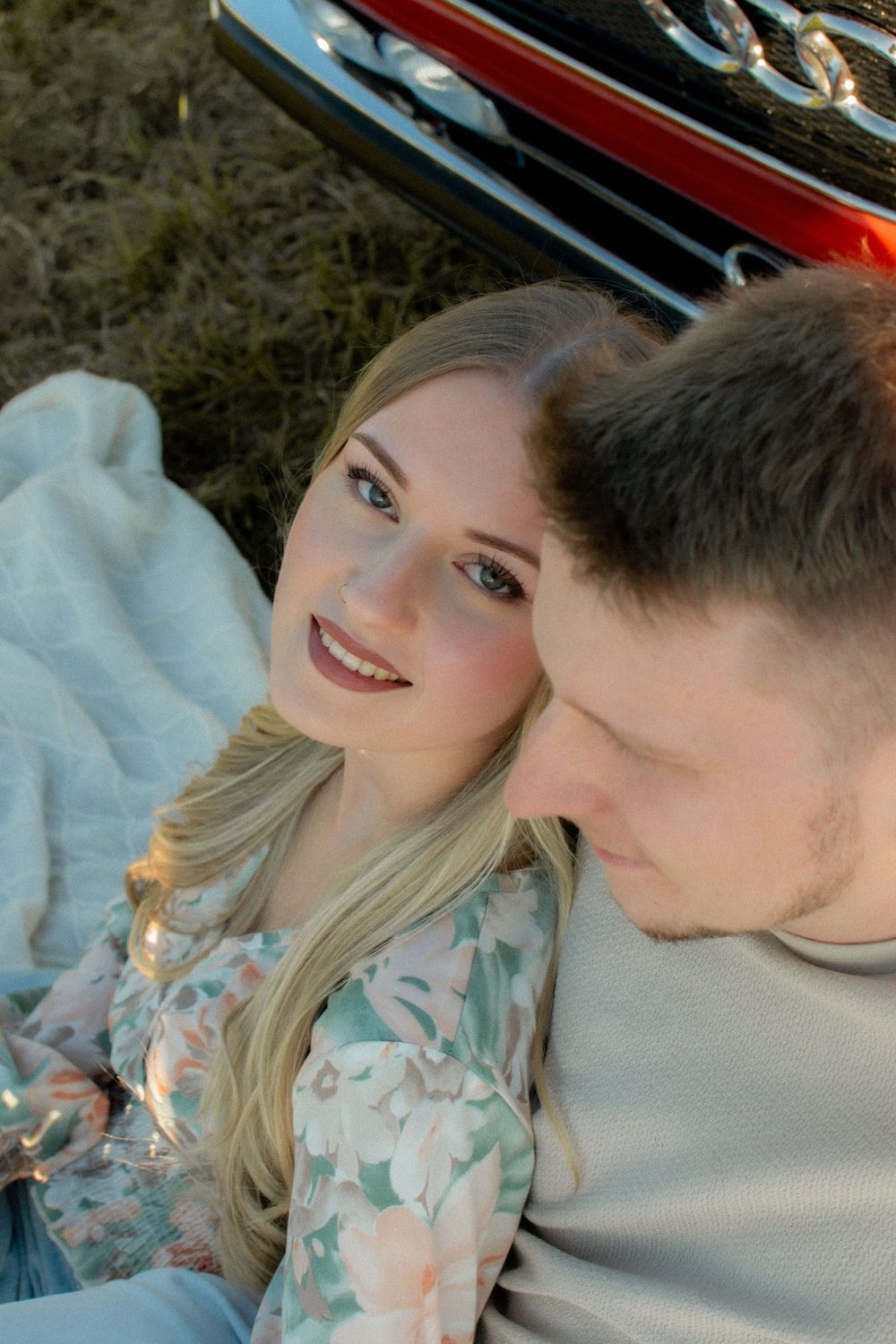 A smiling blonde woman and a man relax together on a blanket outdoors near a vintage red car.