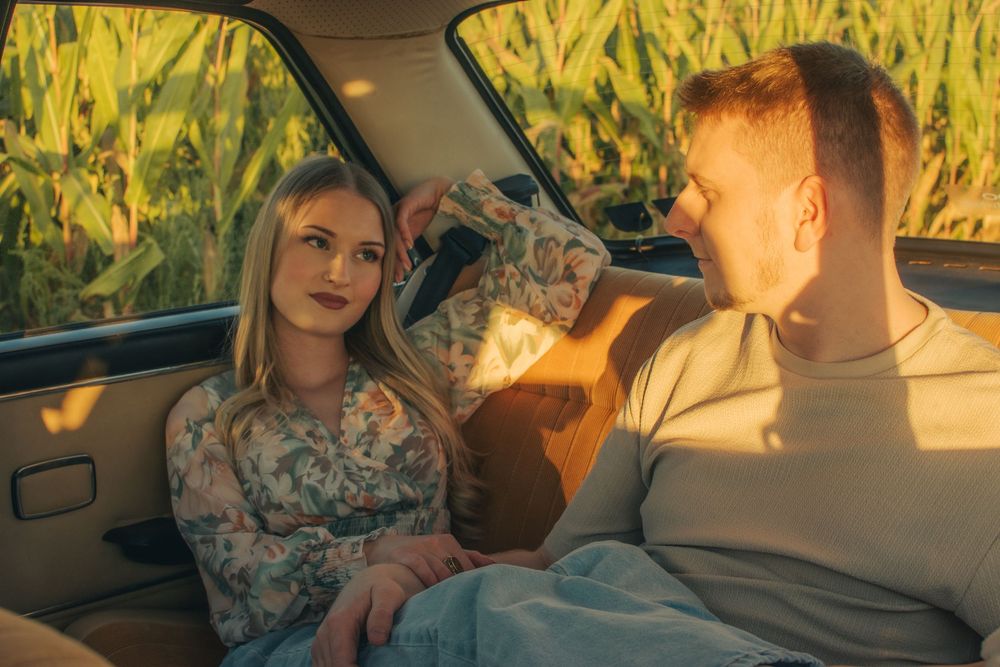Young couple sitting together in a vintage car parked near a cornfield, bathed in warm golden sunlight.