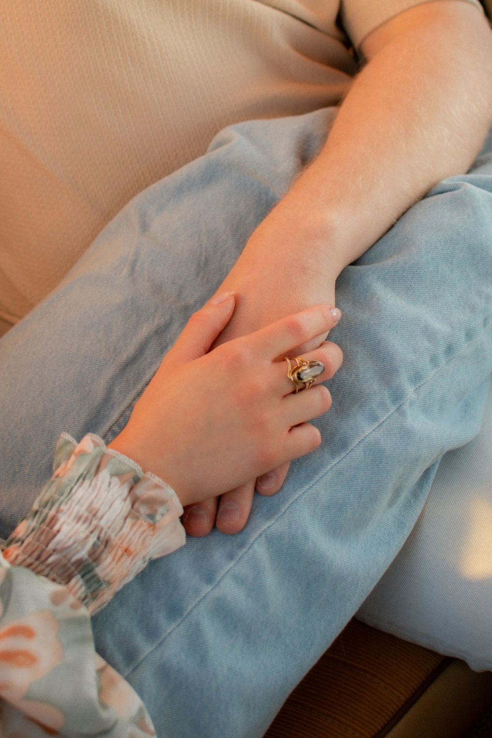Two hands resting together, one wearing a gold gemstone ring, with light blue jeans and a floral sleeve visible.