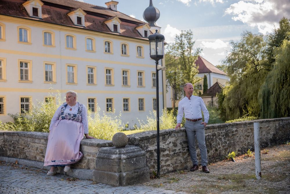 Couple in traditional attire posing near a historic European building and ornate lamppost on a sunny day.