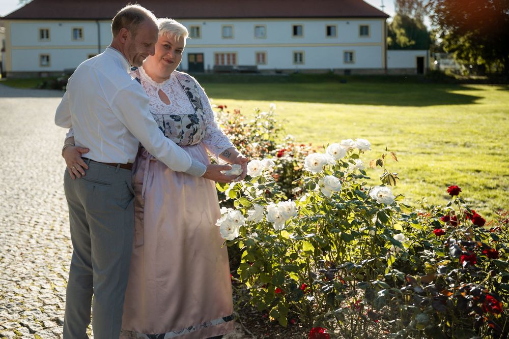 Elderly couple embracing near white rose bushes in a sunny garden with a white building in the background.