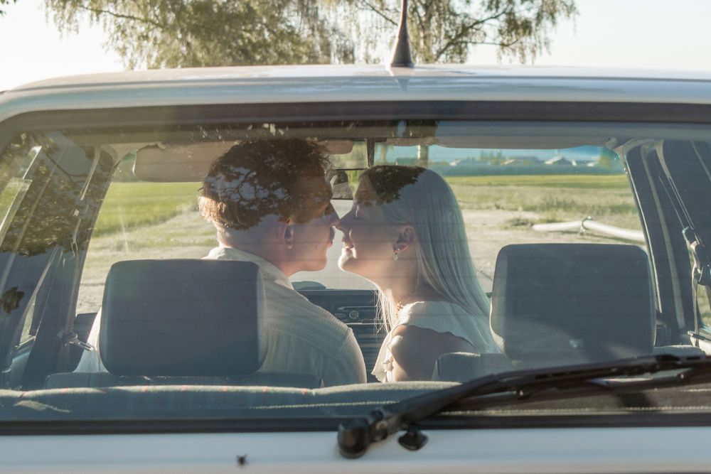 Couple sharing a romantic kiss inside a vintage white car, captured through the windshield at golden hour.