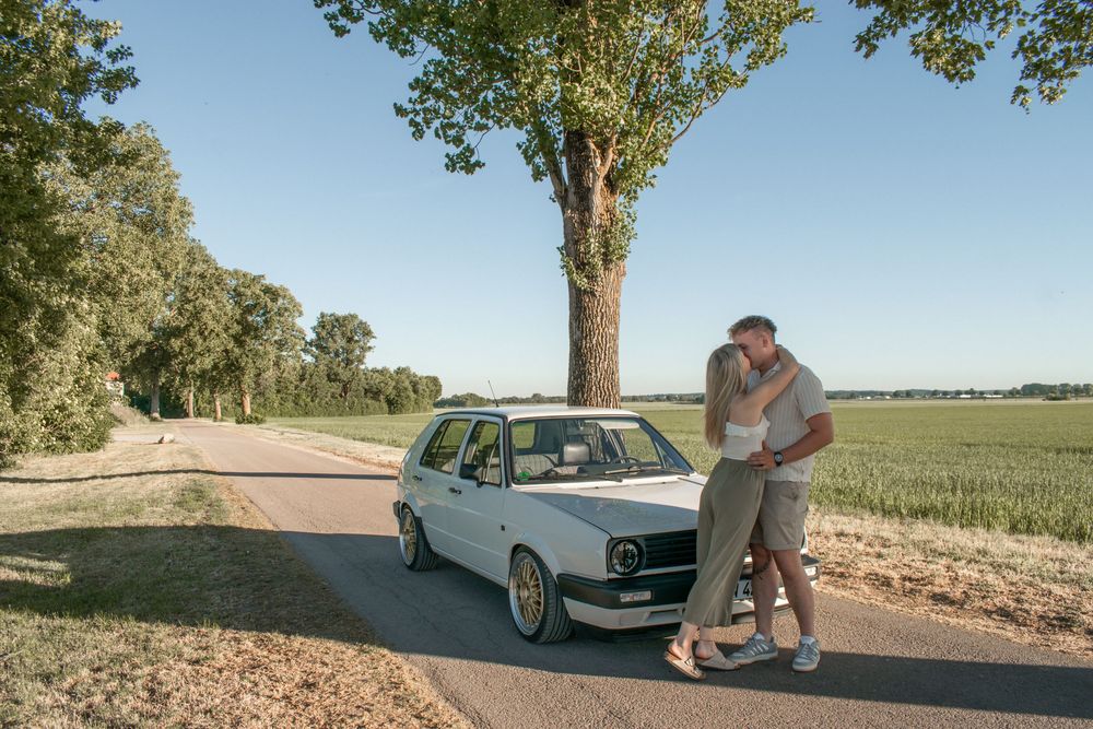 Couple embracing beside a white VW Golf on a rural road lined with trees under a clear blue sky.