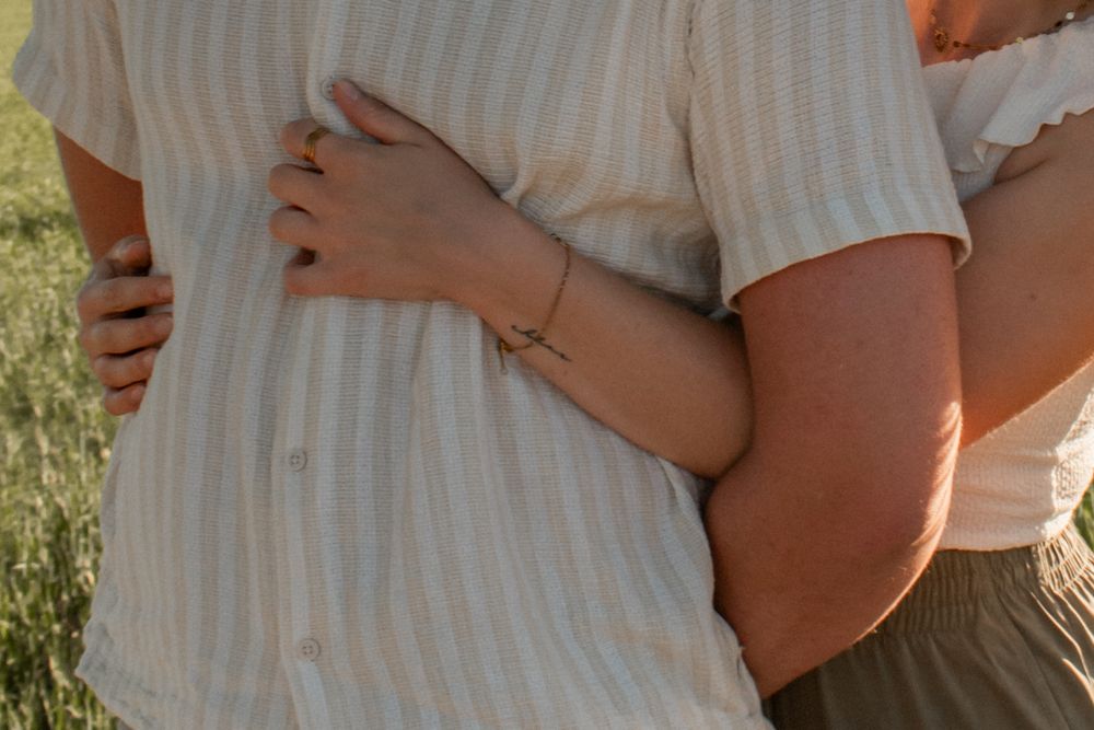 Couple embracing outdoors, woman wearing a delicate gold bracelet, man in striped linen shirt, green field background.