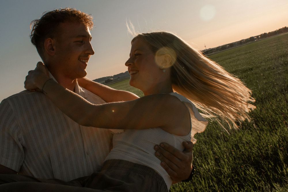 Romantic couple embracing in a sunlit green field at golden hour, smiling joyfully with wind-blown hair.