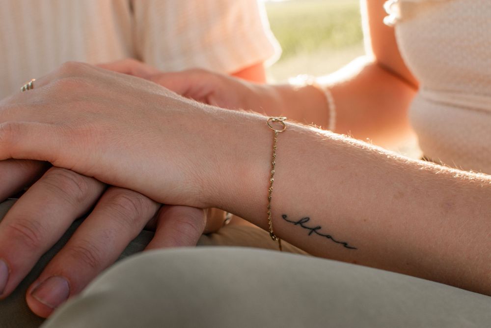Close-up of two hands together, one wearing a delicate gold chain bracelet and the other with a small wrist tattoo.