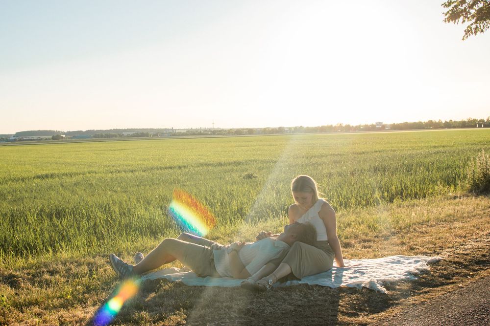 Pregnant woman sitting on blanket in sunny green field at golden hour with lens flare effect.