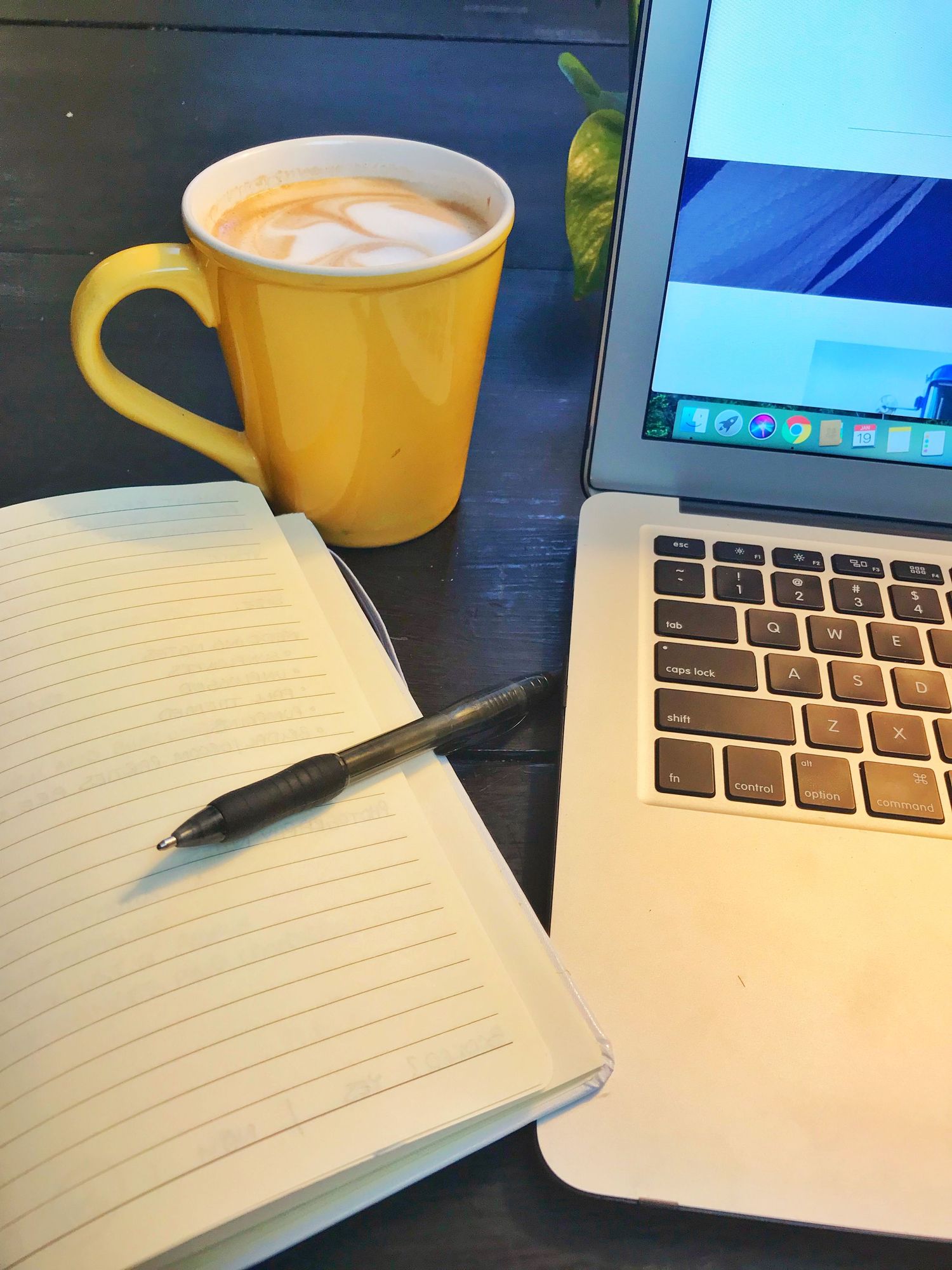 Yellow coffee mug, open notebook with pen, and MacBook laptop on a dark wooden desk workspace.