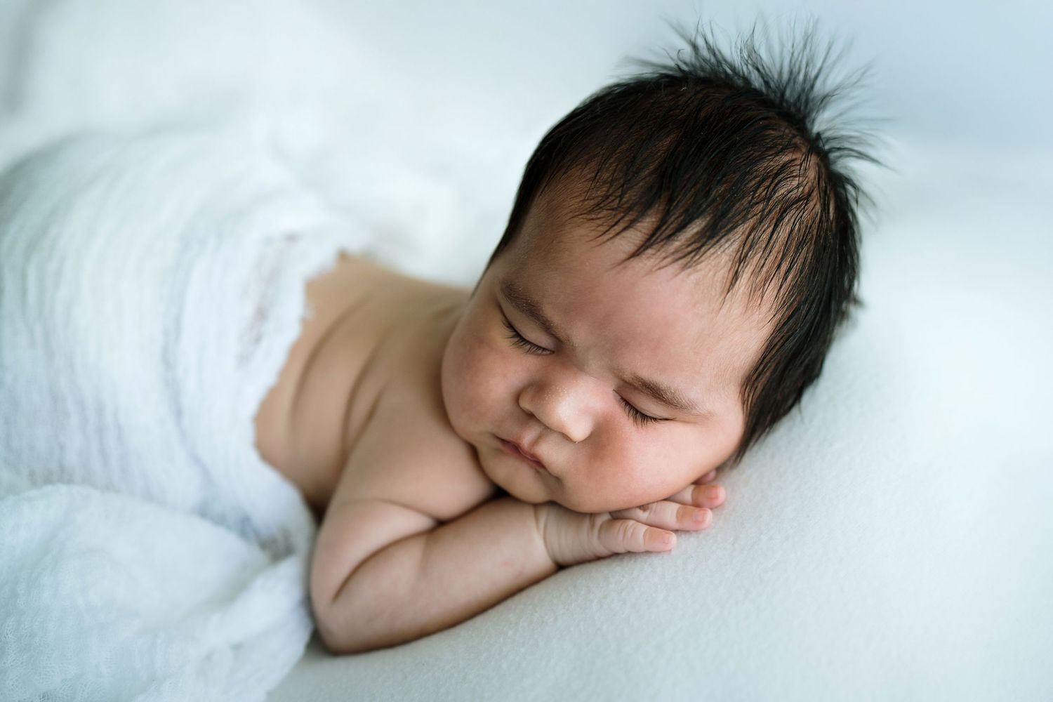 Peaceful sleeping newborn with dark hair rests on white blanket.