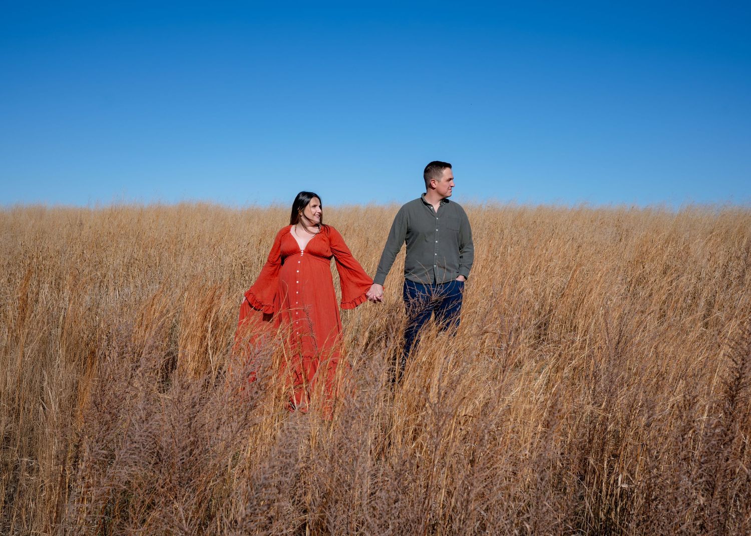 A maternity session with husband and wife holding hands side by side in a tall golden grass field looking off in the distance.