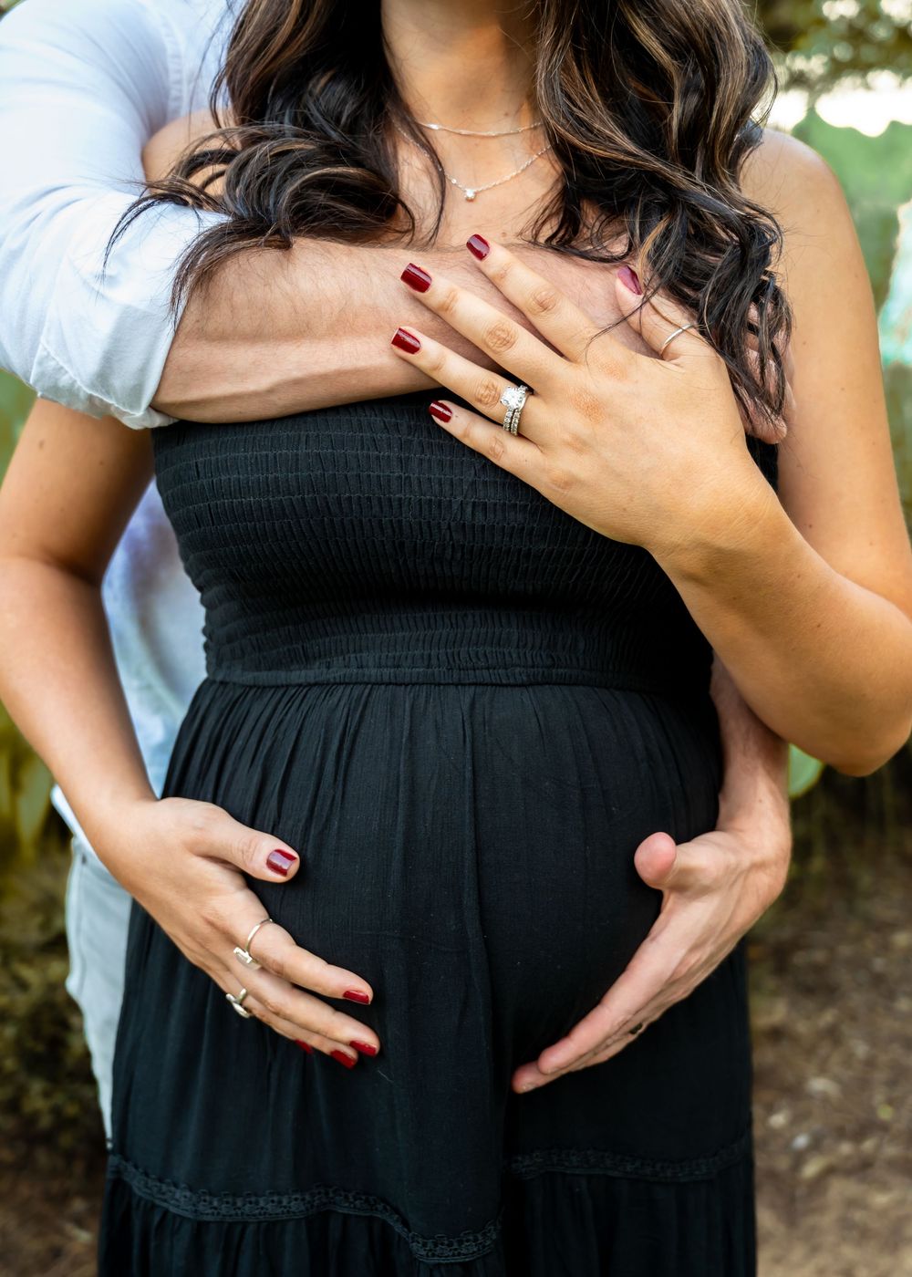 A couple cradles a pregnant belly outdoors, showing off wedding rings and red manicured nails.