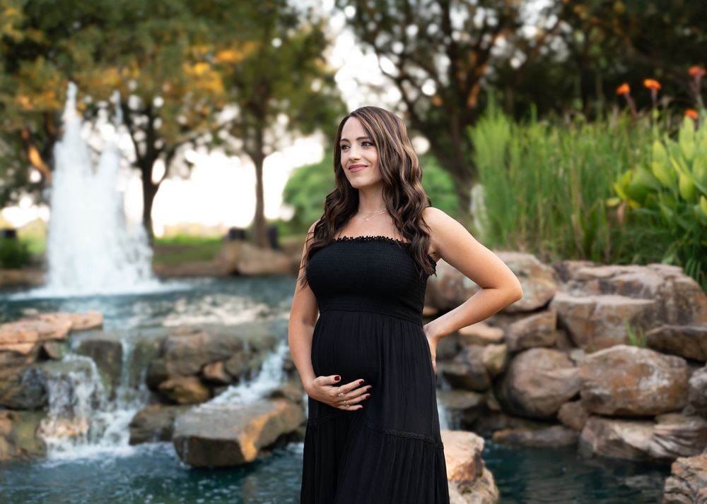 Pregnant woman in black maxi dress poses by fountain during golden hour maternity shoot.