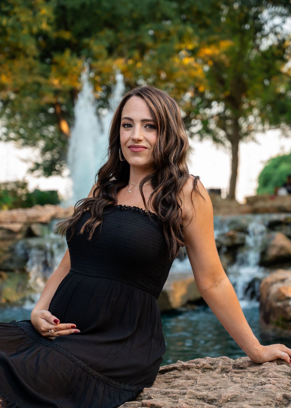 Smiling pregnant woman in black dress sits on rocks near fountain for maternity photos.