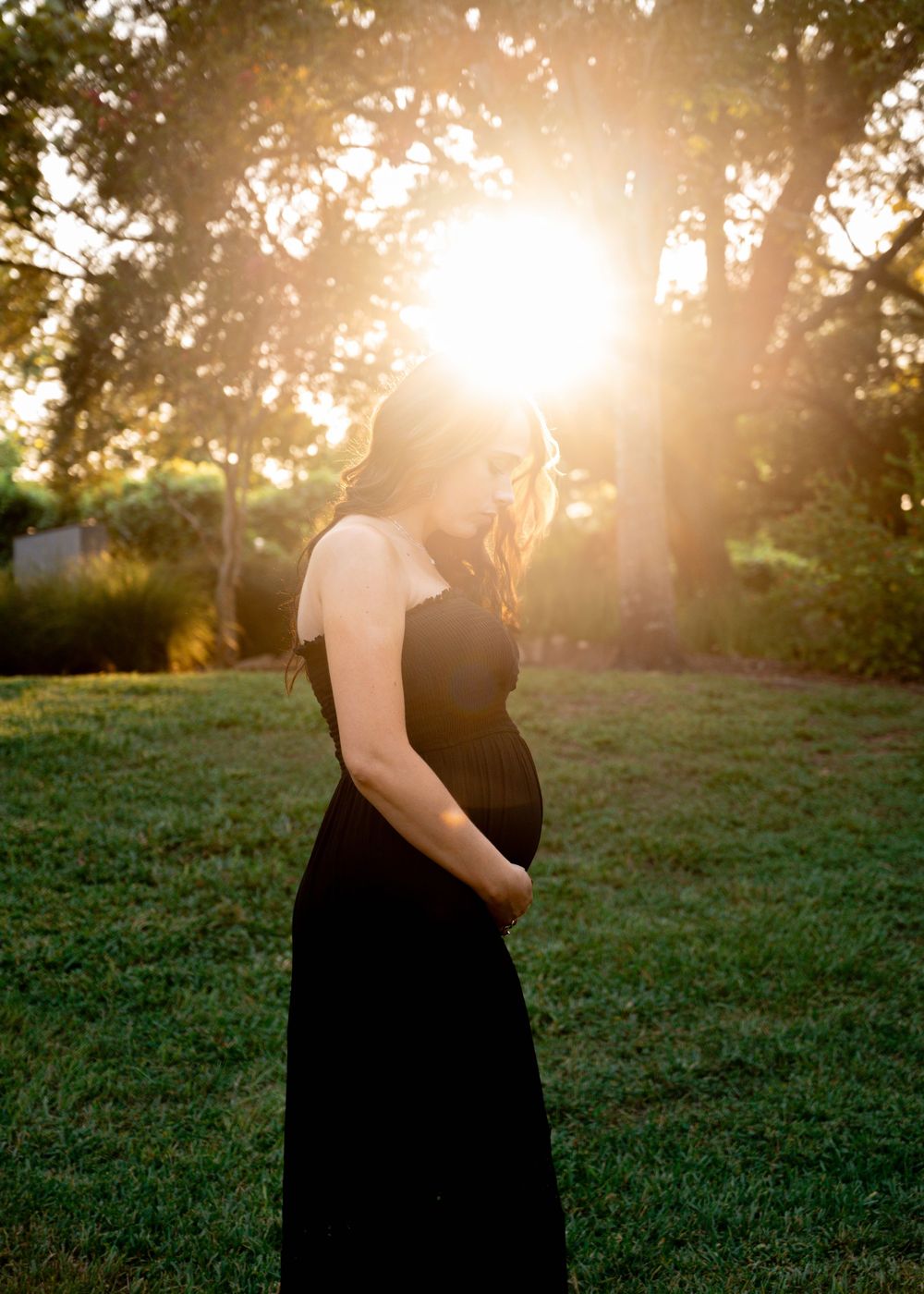 Pregnant woman in black dress cradles baby bump during stunning golden sunset maternity session.