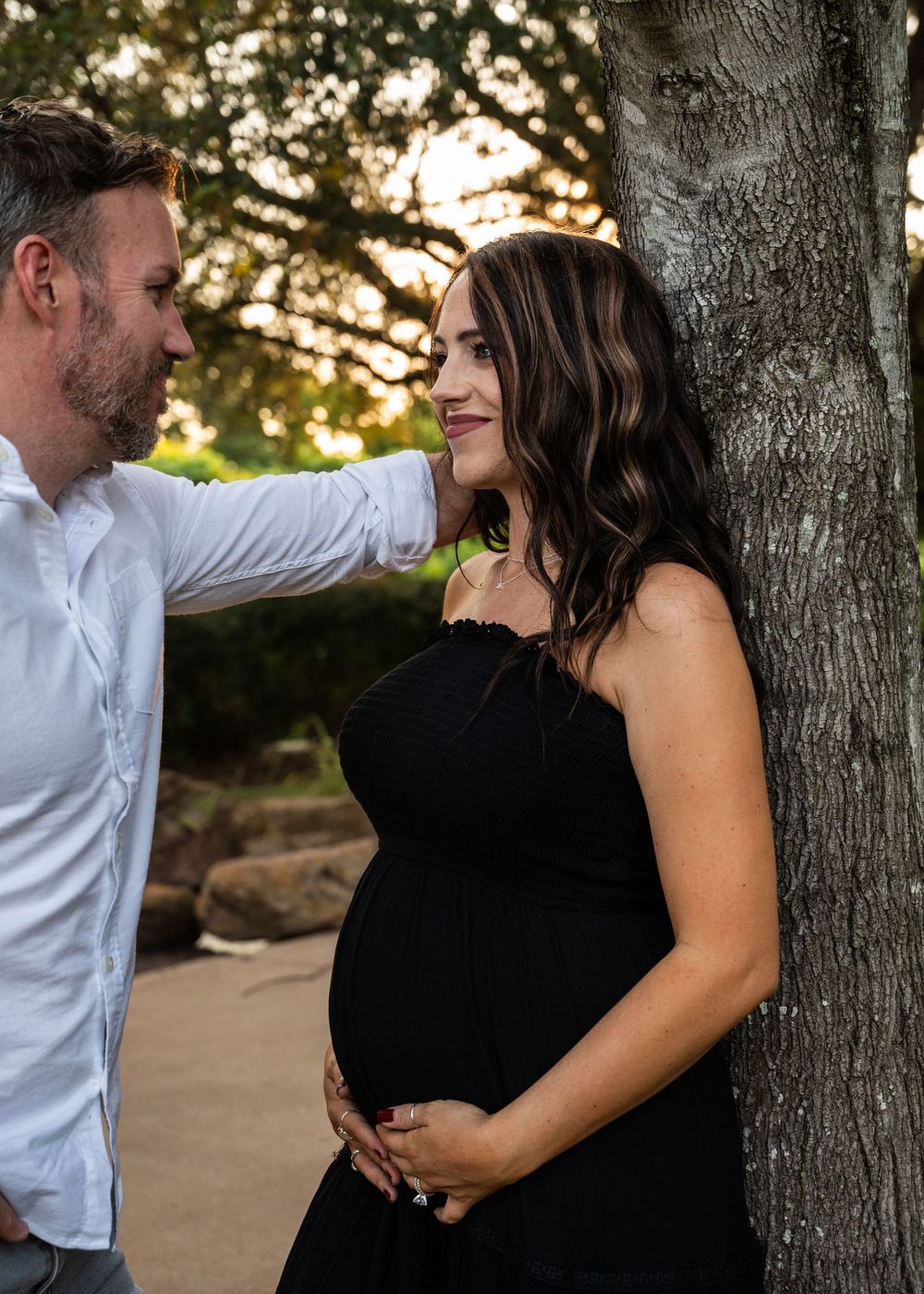 Couple shares tender moment by tree during outdoor maternity photography session at sunset.