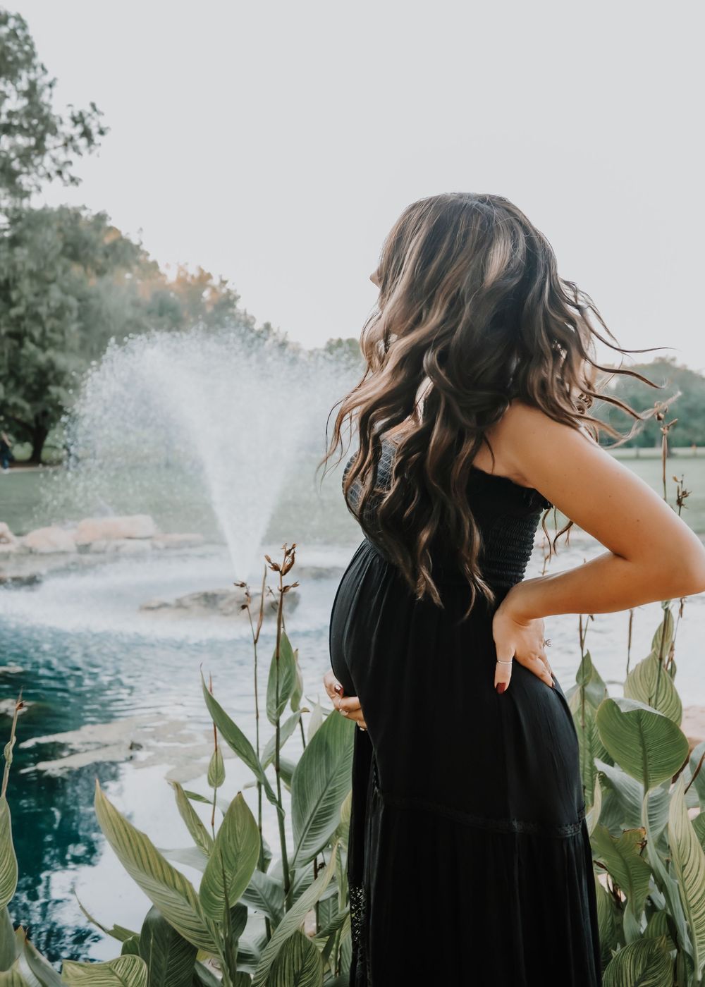 Pregnant woman in black dress gazes at fountain during outdoor maternity photo session.