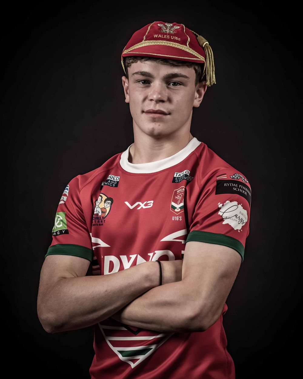 Rugby player in red jersey and traditional uniform cap poses against dark backdrop.