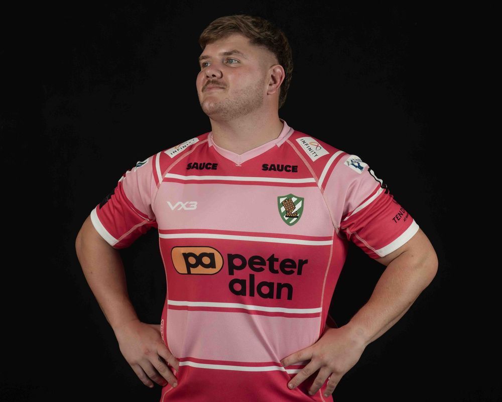 Rugby players wearing pink jerseys with advertising logos pose against dark background.