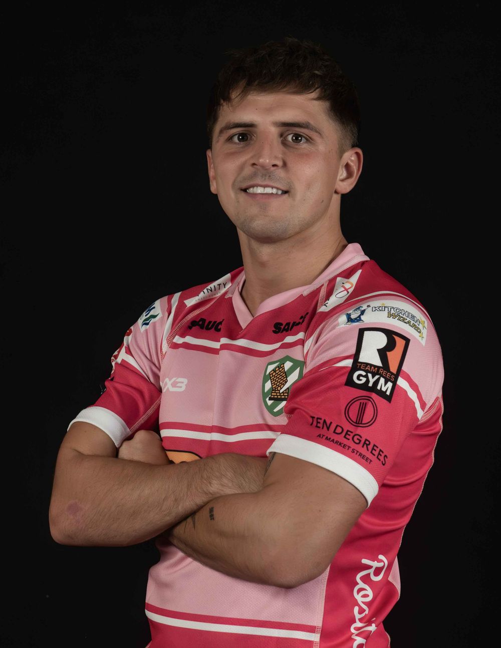Rugby player poses in pink and white striped uniform against dark background with folded arms.