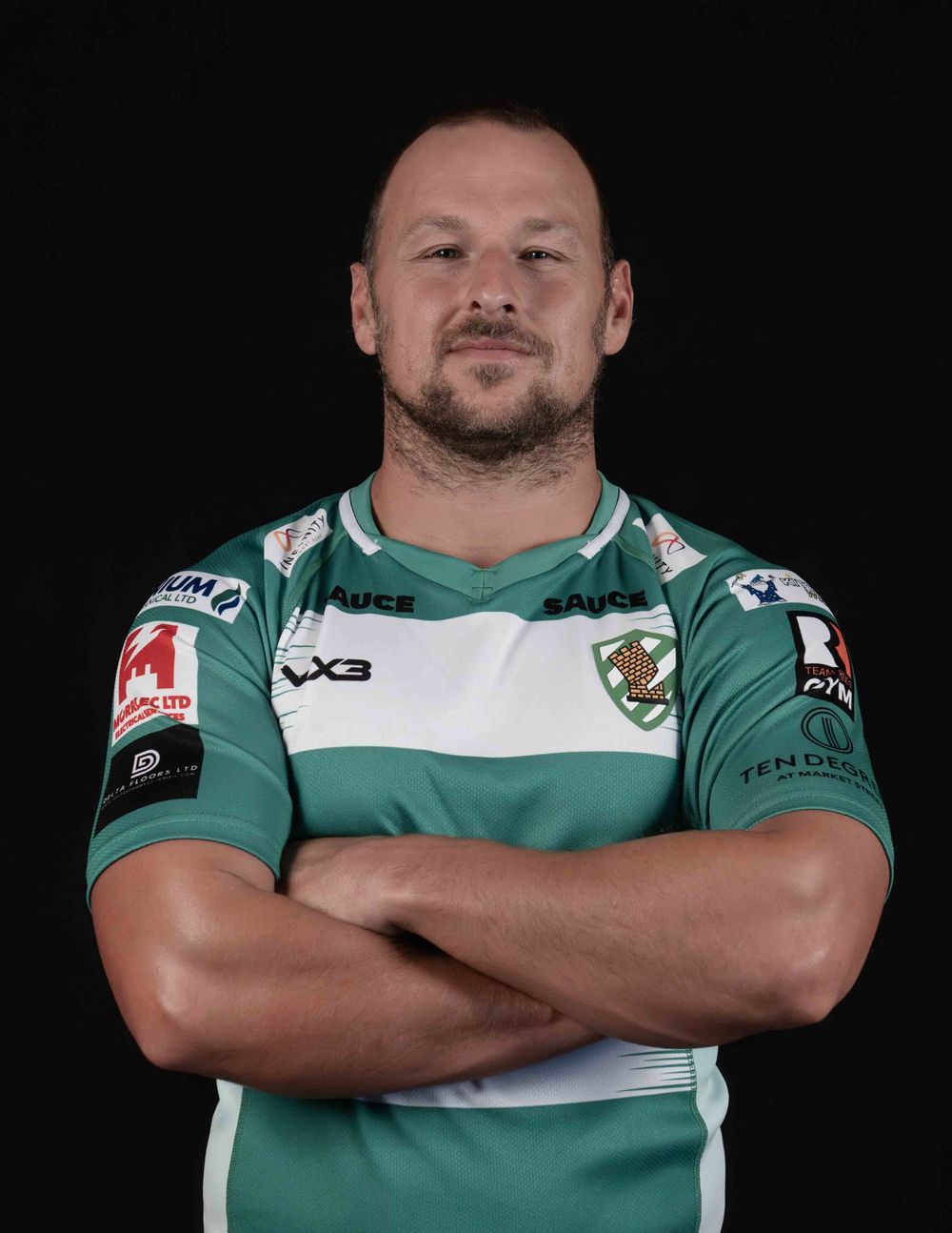 Rugby player shown in green and white uniform stands with crossed arms against black backdrop.