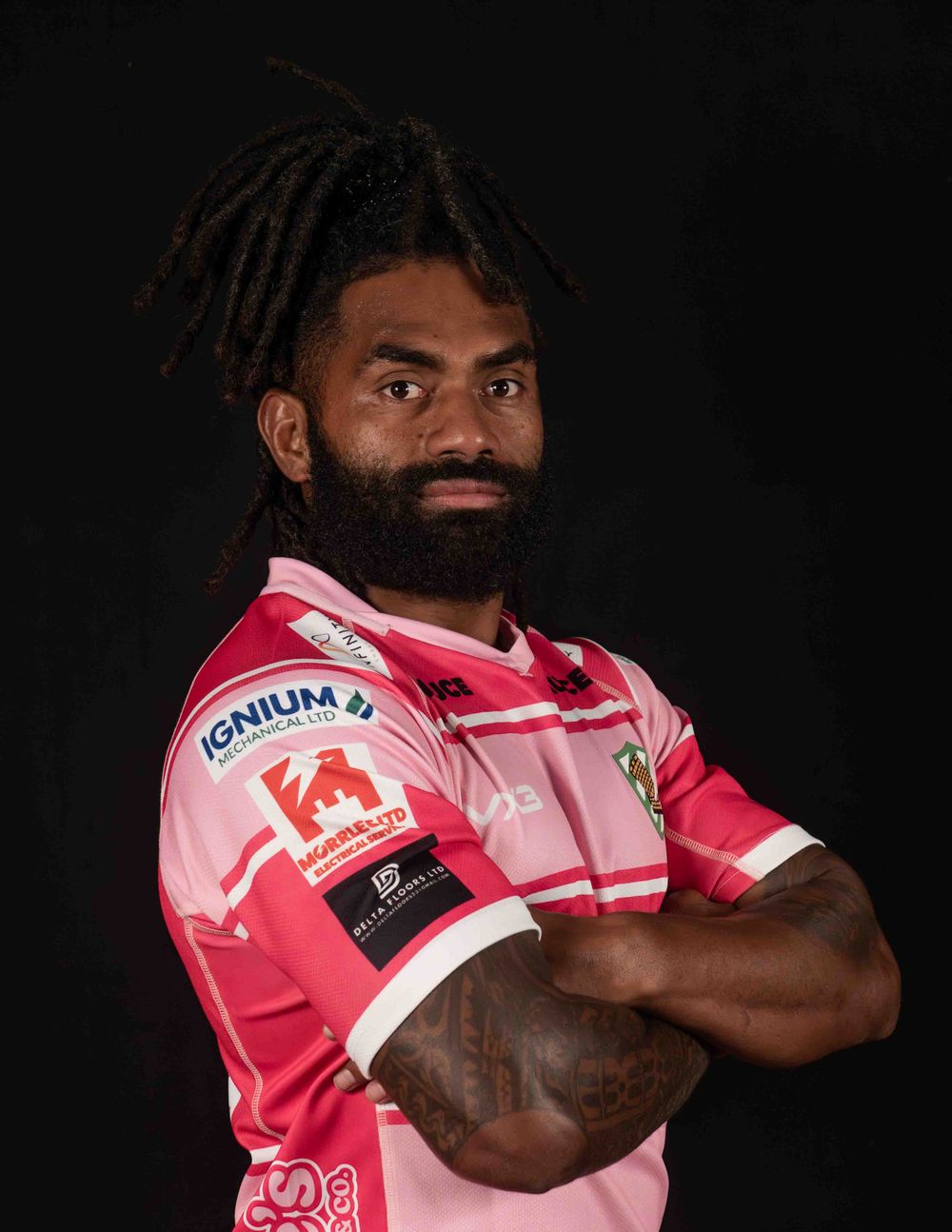 Rugby player wearing pink jersey against dark background poses with crossed arms.