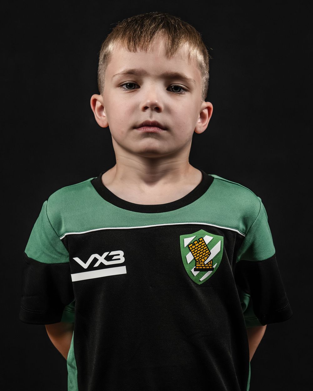 A youth soccer player wearing a black and green uniform poses against a dark background for a team photo.