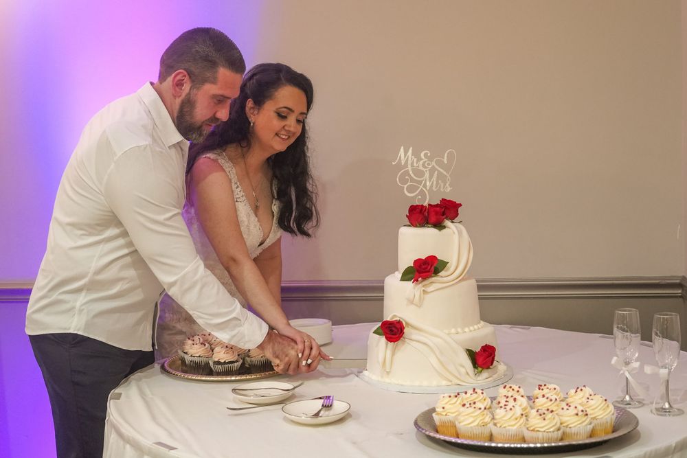 Wedding couple cuts their white tiered cake decorated with red roses at their reception.