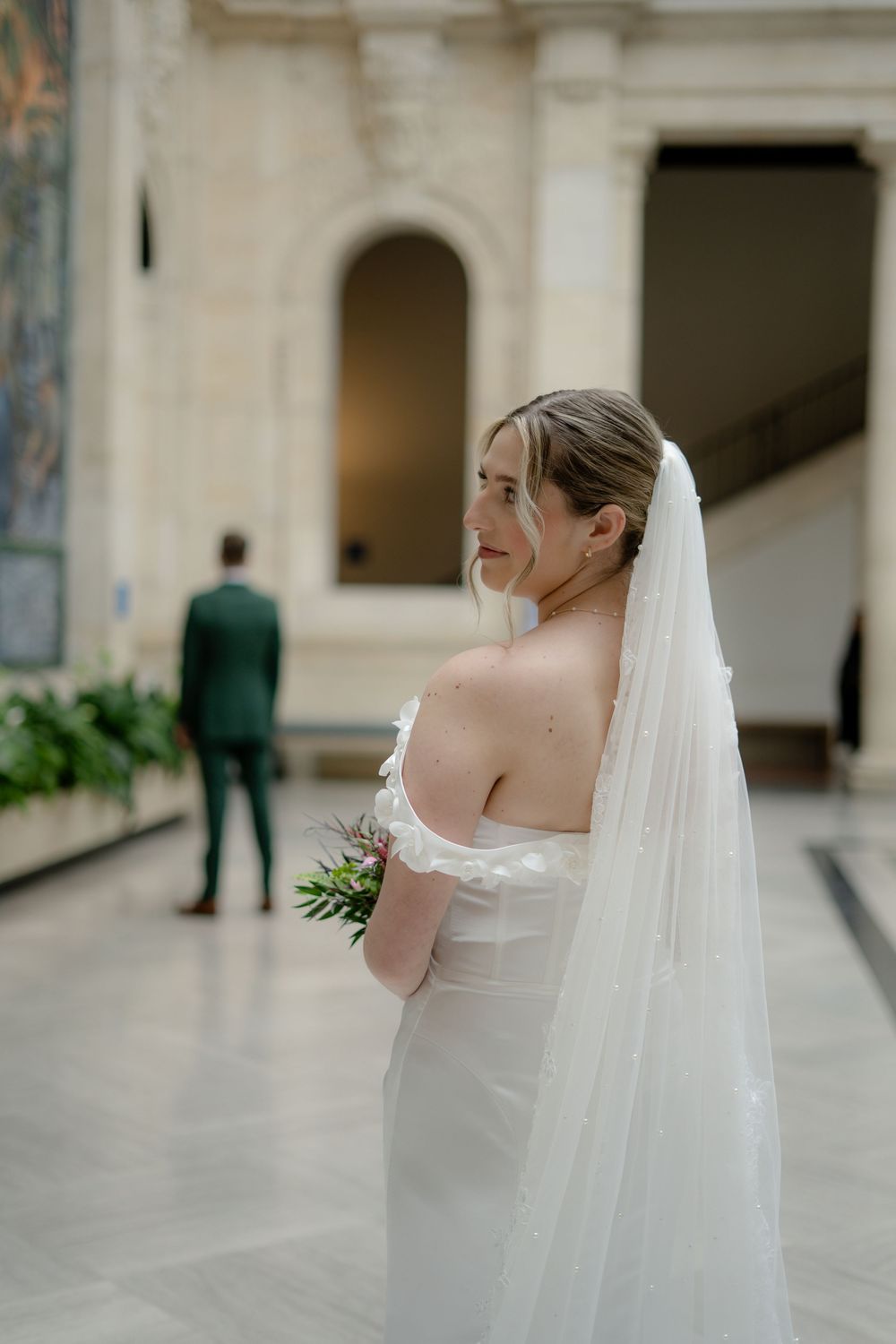 Elegant bride in white dress and flowing veil stands in marble hallway.