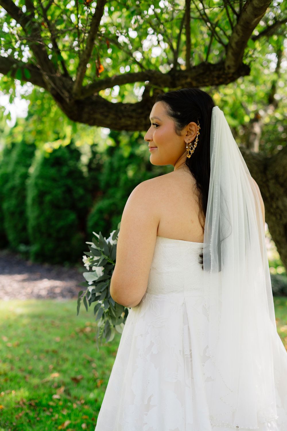 A bride in a strapless wedding gown poses under green trees with a flowing veil.