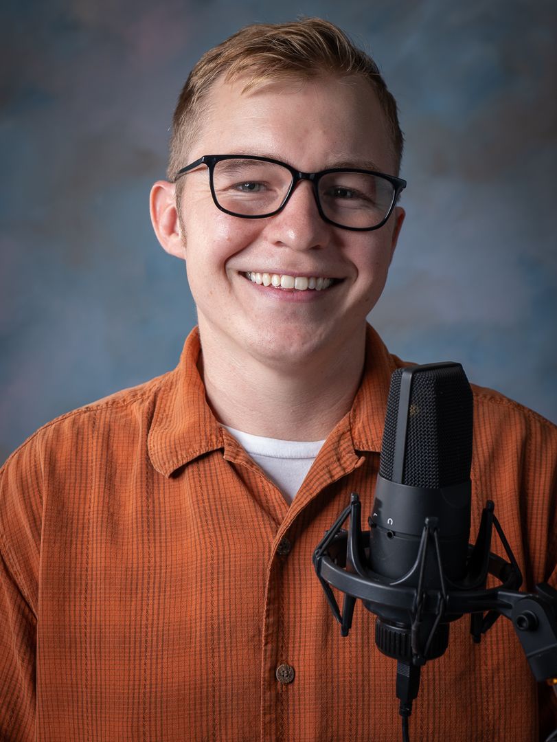 Portrait of a podcaster wearing glasses and orange shirt in front of condenser microphone.