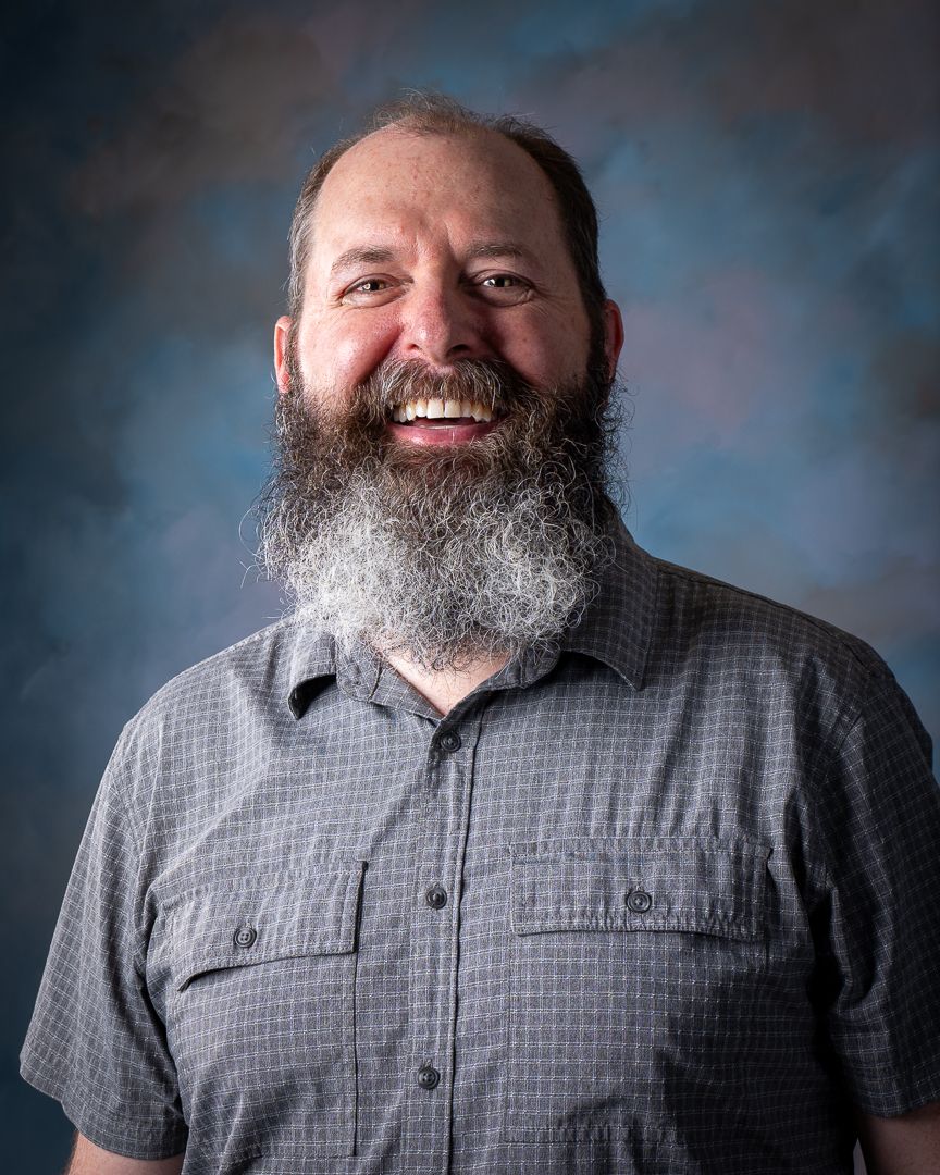 Portrait of a bearded person in gray shirt against blue backdrop smiling warmly.