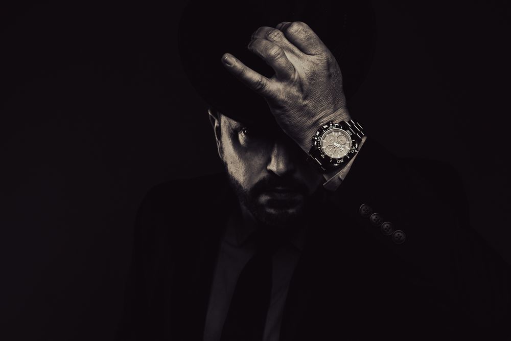 Dramatic low-key portrait showing a person's dark silhouette with a watch-adorned hand raised against a black background.
