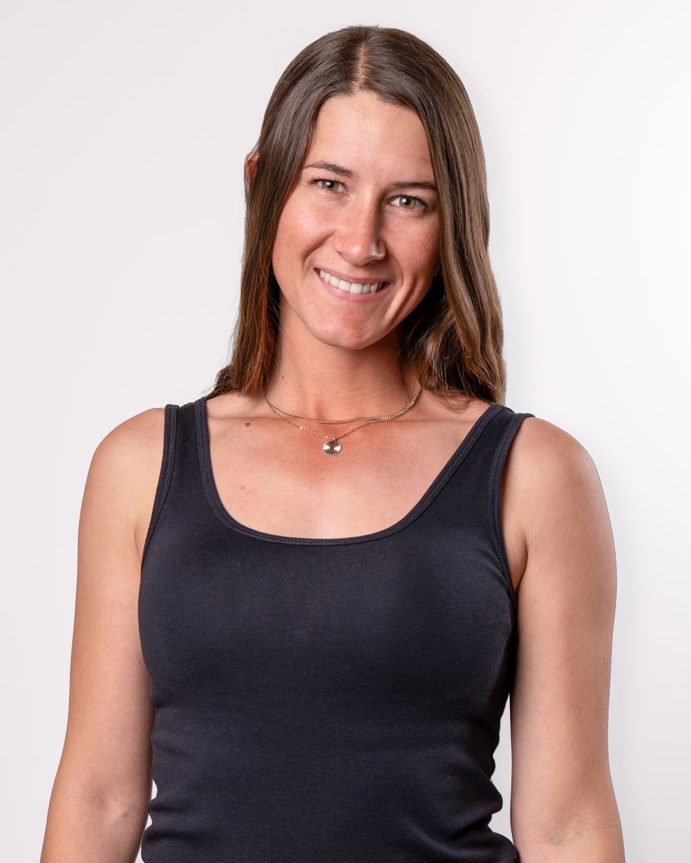 Studio portrait with warm expression wearing black tank top and dainty necklace against light background.