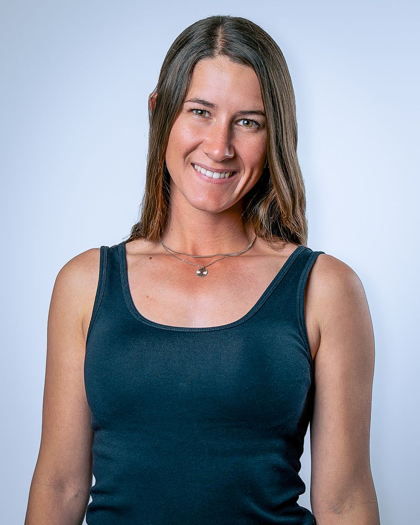 Professional headshot of a person in a navy tank top with a delicate necklace against a light gray background.