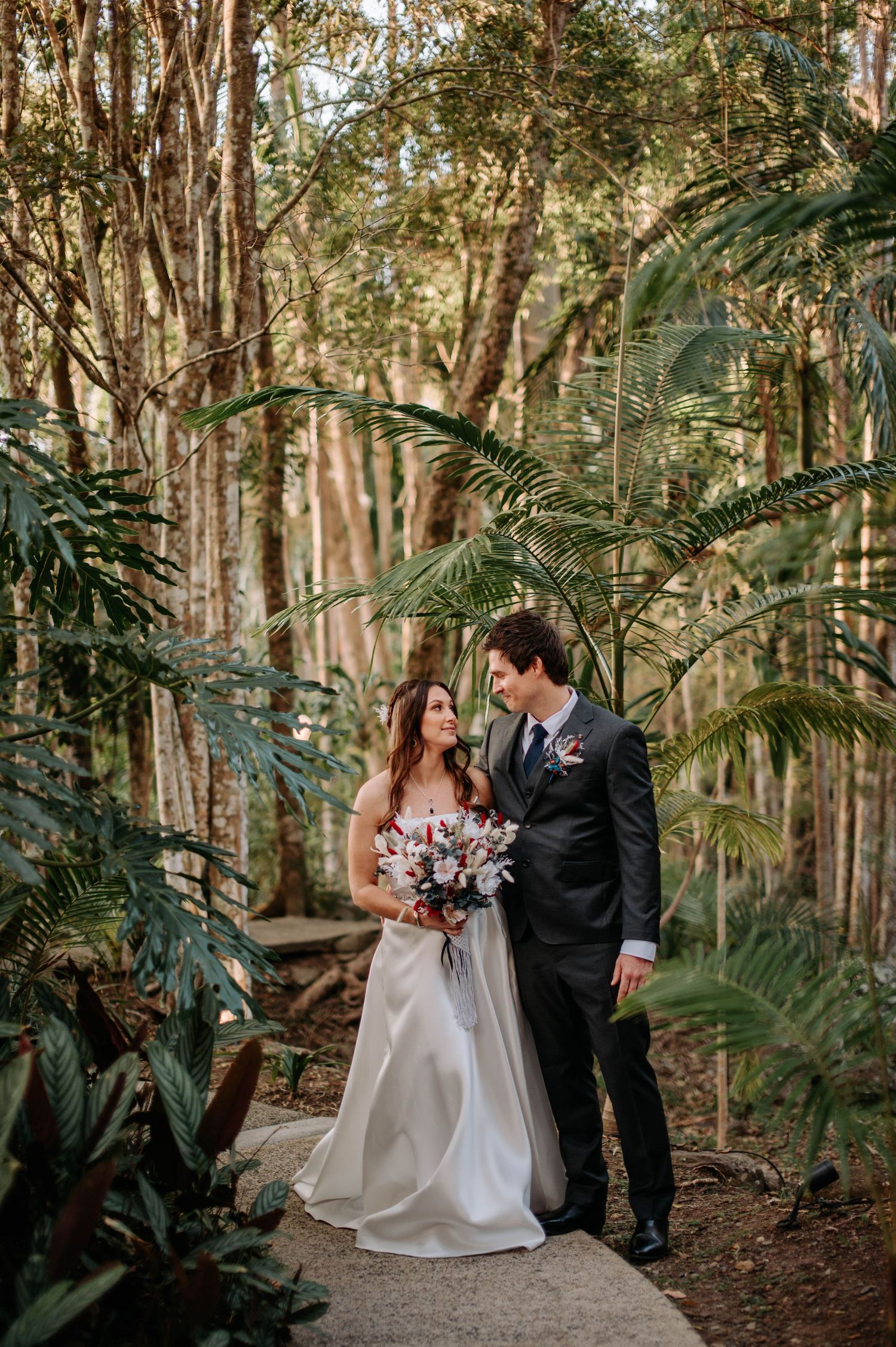 Couple shares intimate moment on garden path surrounded by tropical foliage and tall trees.