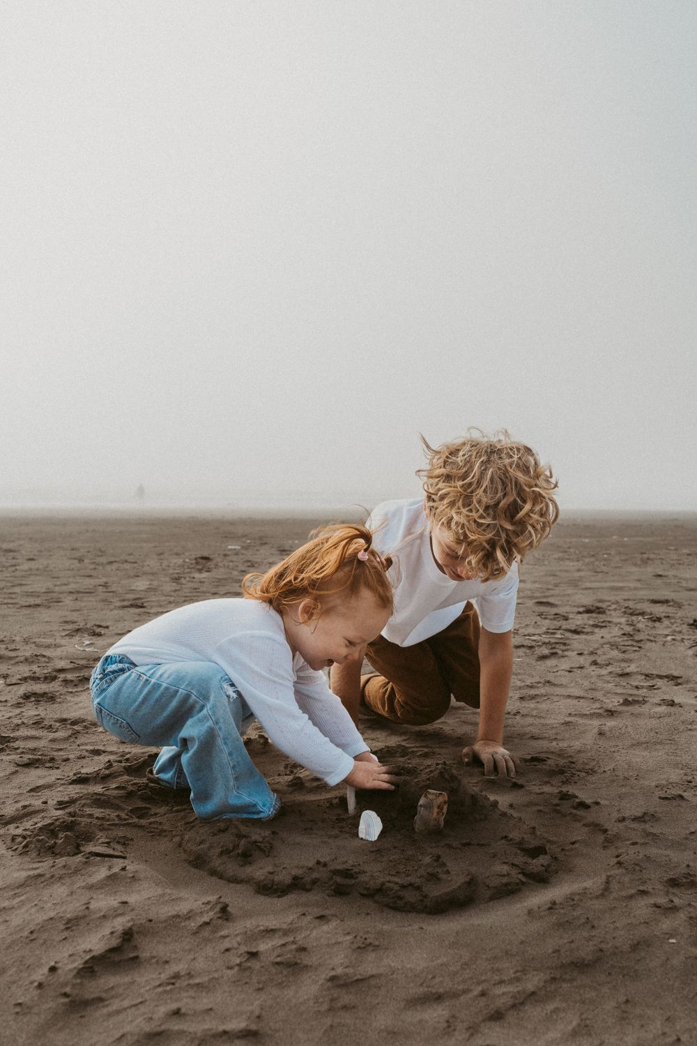 Two children playing together in the sand on a misty beach at dusk.