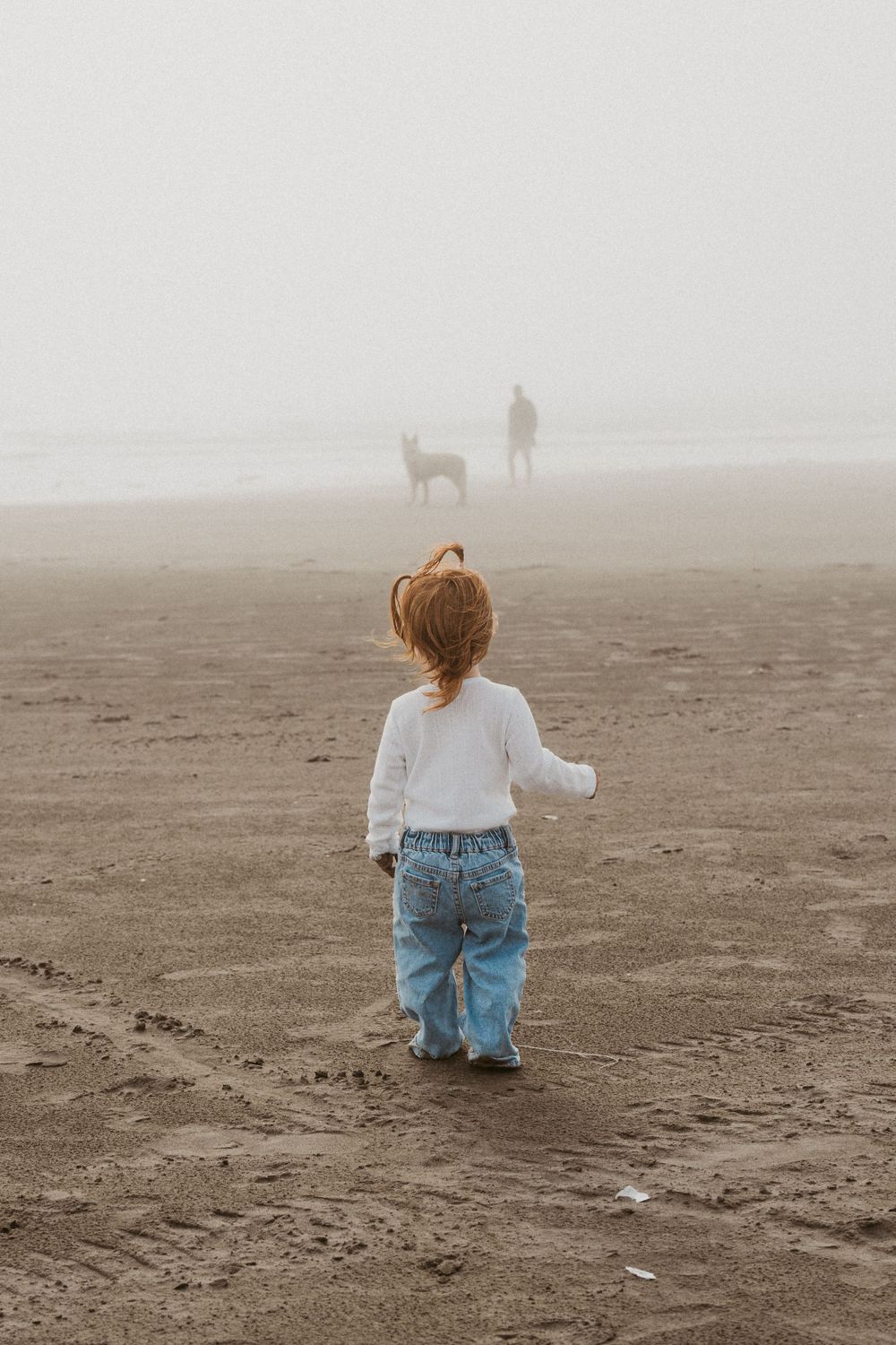 Child in jeans and white sweater watching figures in fog on beach.