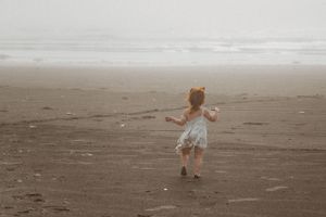 Small child in white dress twirling joyfully on foggy beach.