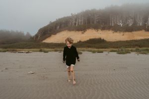 Person walking barefoot on a foggy beach with cliffs and trees in background.