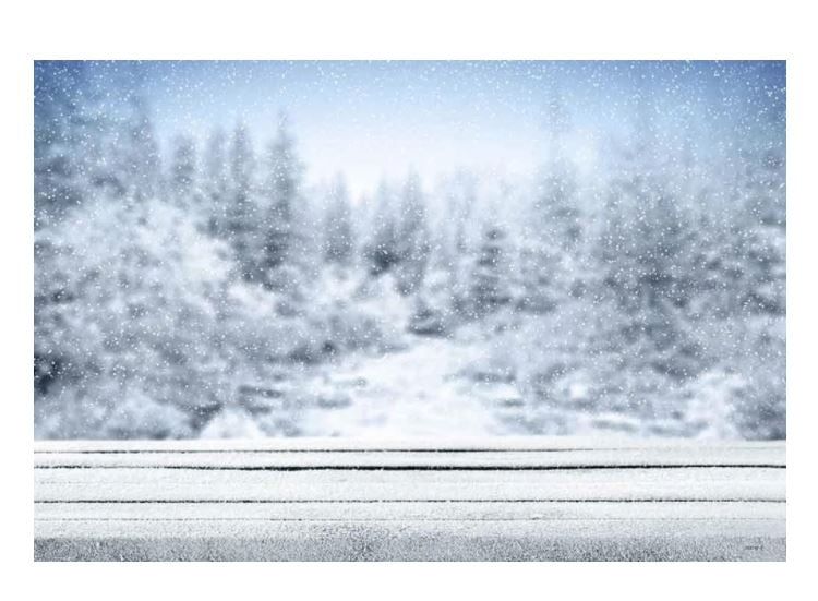 Snow-covered winter forest with falling snowflakes and frosted pine trees behind a rustic wooden table dusted with snow.