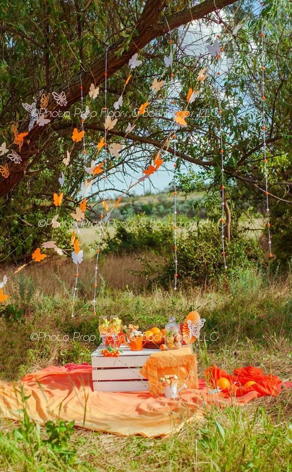 Outdoor picnic setup with orange butterfly garlands hanging from tree branches over a white crate and colorful blankets.