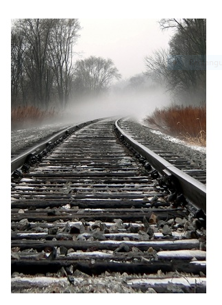 Snow-dusted railroad tracks curve into misty fog, flanked by bare winter trees in a serene rural landscape.