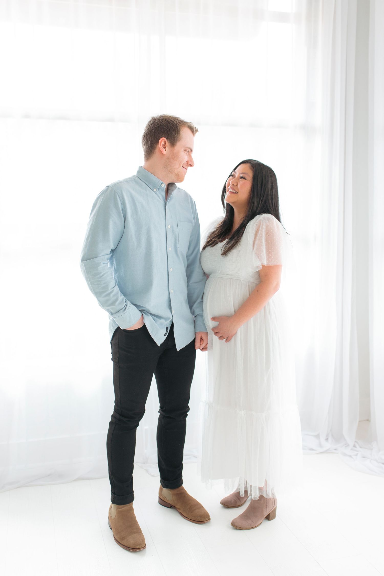 Expecting couple sharing a joyful moment together against white curtains in a bright minimal setting.