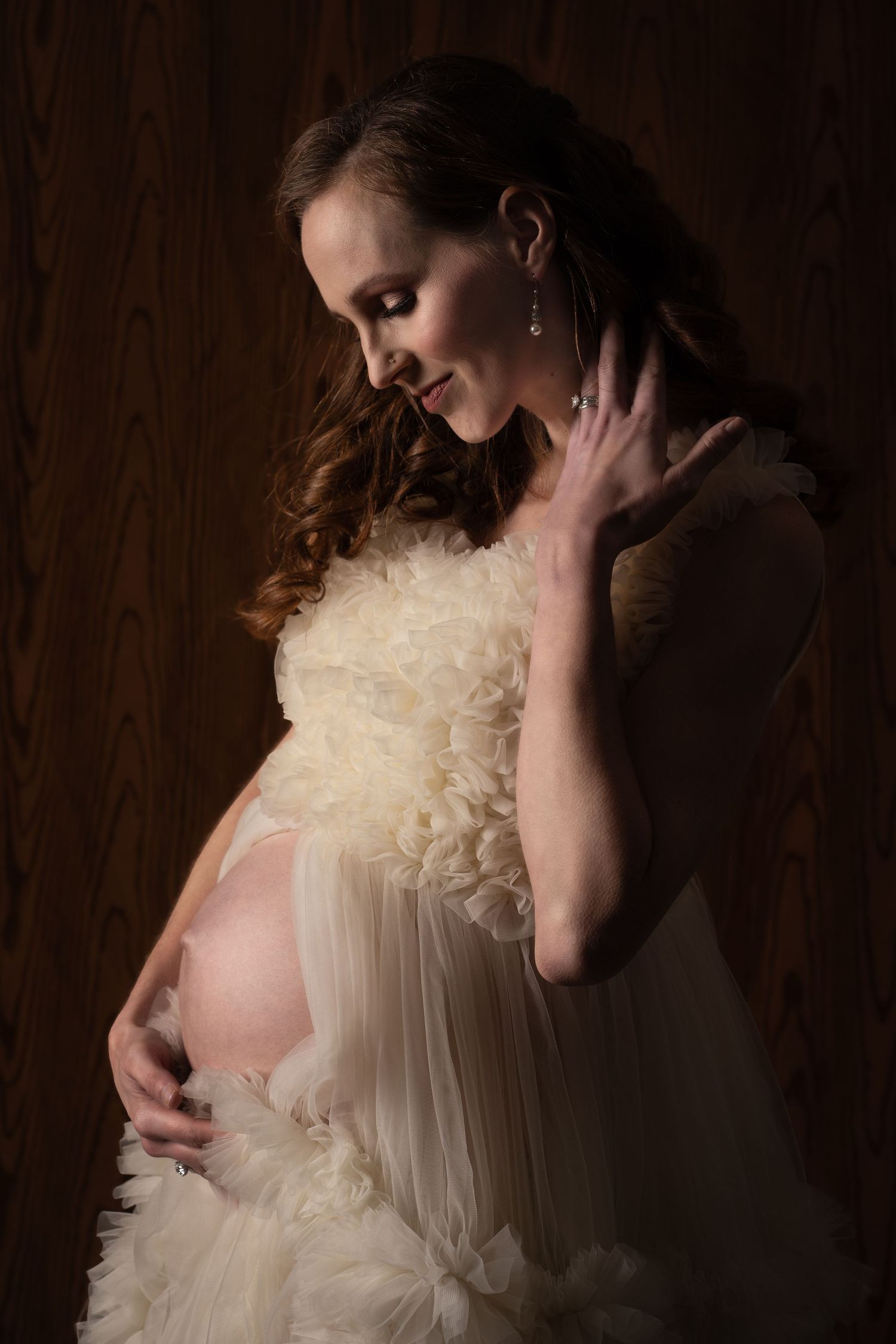 Dramatic side-lit maternity portrait in cream ruffled gown against dark wooden wall.