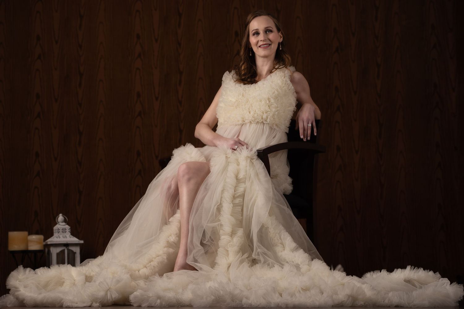 Expectant mother in luxurious cream gown poses on chair with flowing fabric around her.
