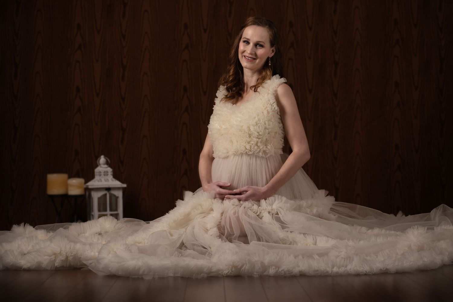 Pregnant woman in elegant cream ruffled gown sitting gracefully on floor with lantern decor in professional maternity photo.