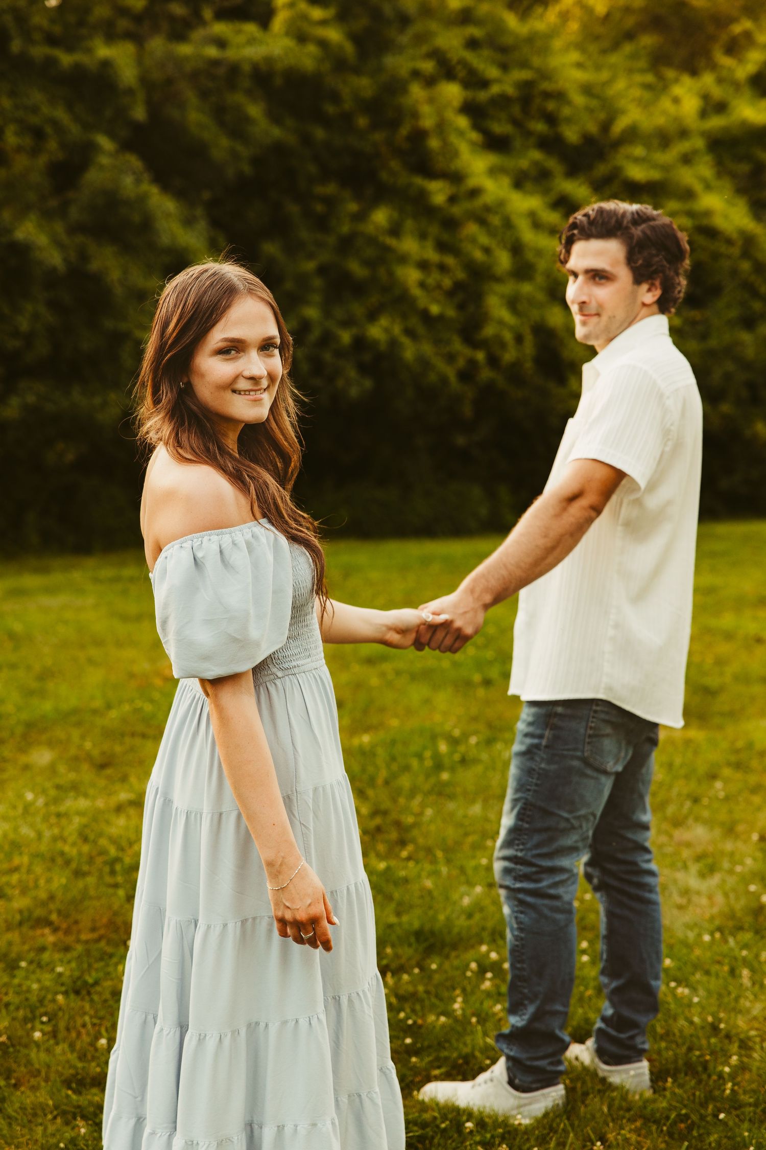 A romantic couple holds hands in a grassy field during golden hour, with her in a blue off-shoulder dress and him in white.