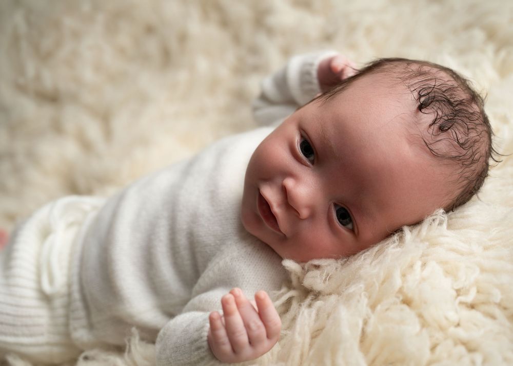 Newborn baby in white outfit lying on fluffy cream rug, looking up with dark hair and bright eyes.