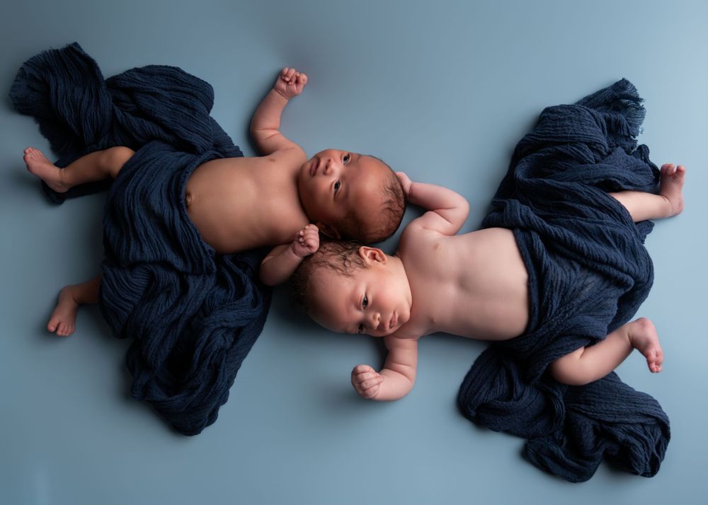 Two newborn twins sleeping side by side wrapped in dark navy blue fabric on a blue background.