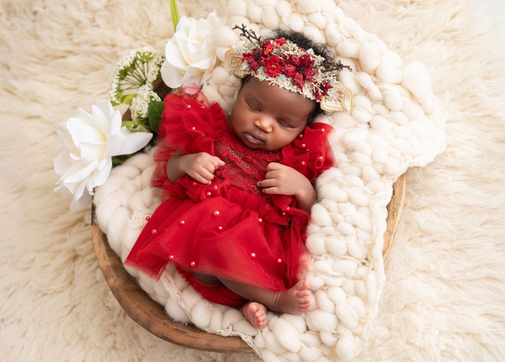 A newborn baby wearing a red velvet dress lies peacefully on a cream-colored blanket during a professional photo session.
